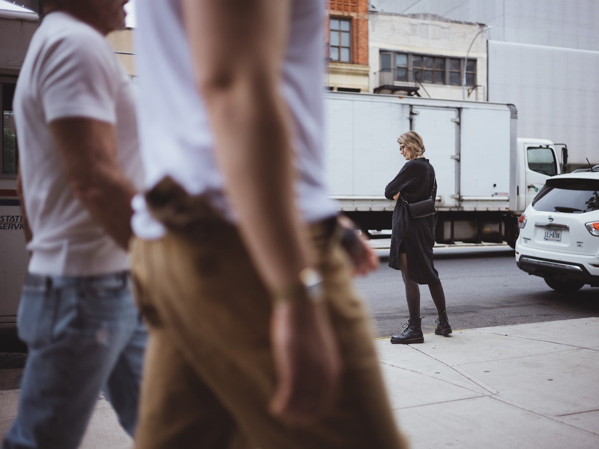 A woman in black stands on a city sidewalk.
