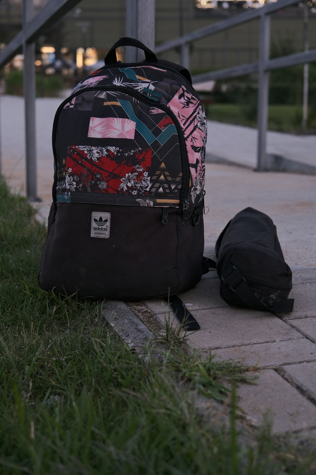 A patterned backpack and a small black cylindrical bag on a paved walkway.